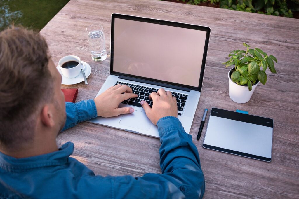 Man typing on laptop outfoors with coffee, water and plant beside him.