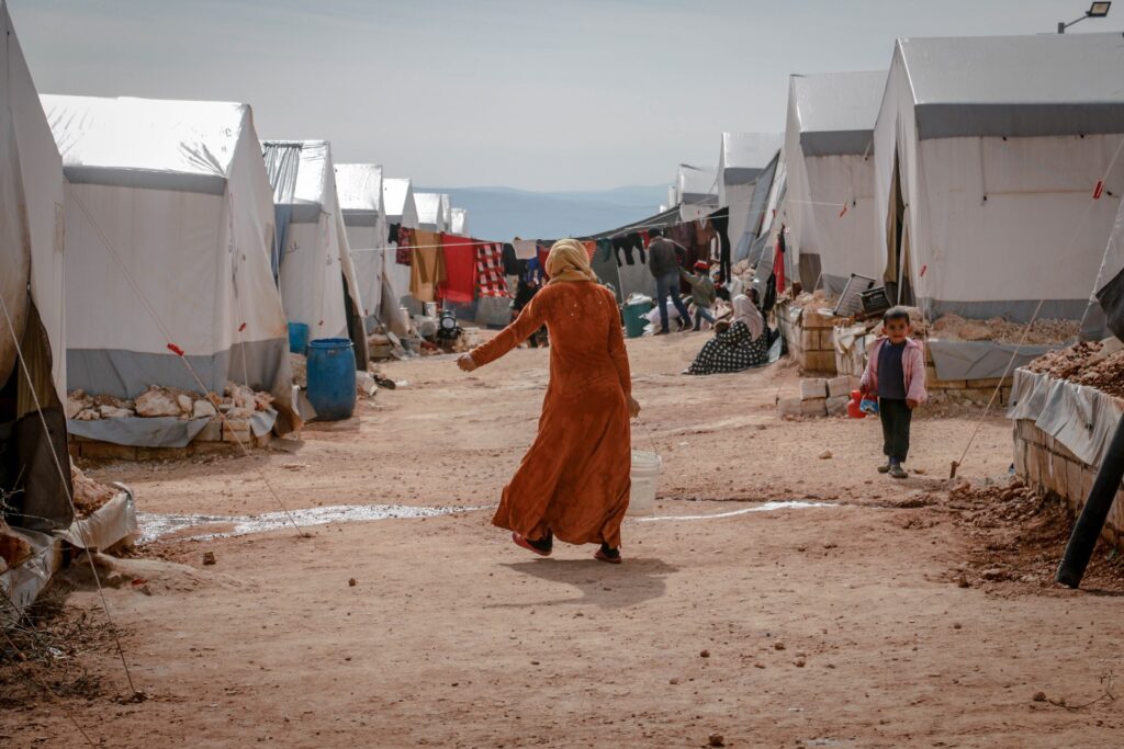 A woman standing in the middle of a dirt road which is in between refugee tents. This represents VR is the ultimate empathy machine by bringing people to the refugee situation.