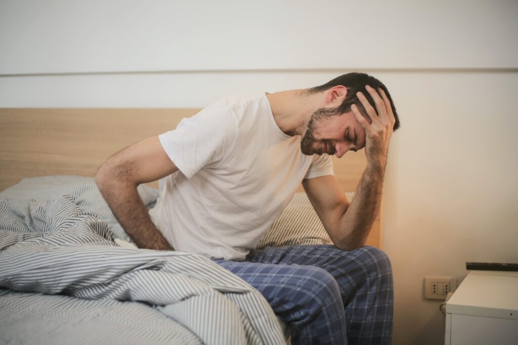 A man sits on the side of a bed with his hand on his head. He appears to be in pain.