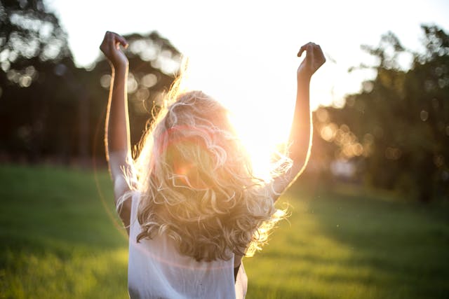 Image of girl with blonde hair standing in forest with arms up in front of sun.