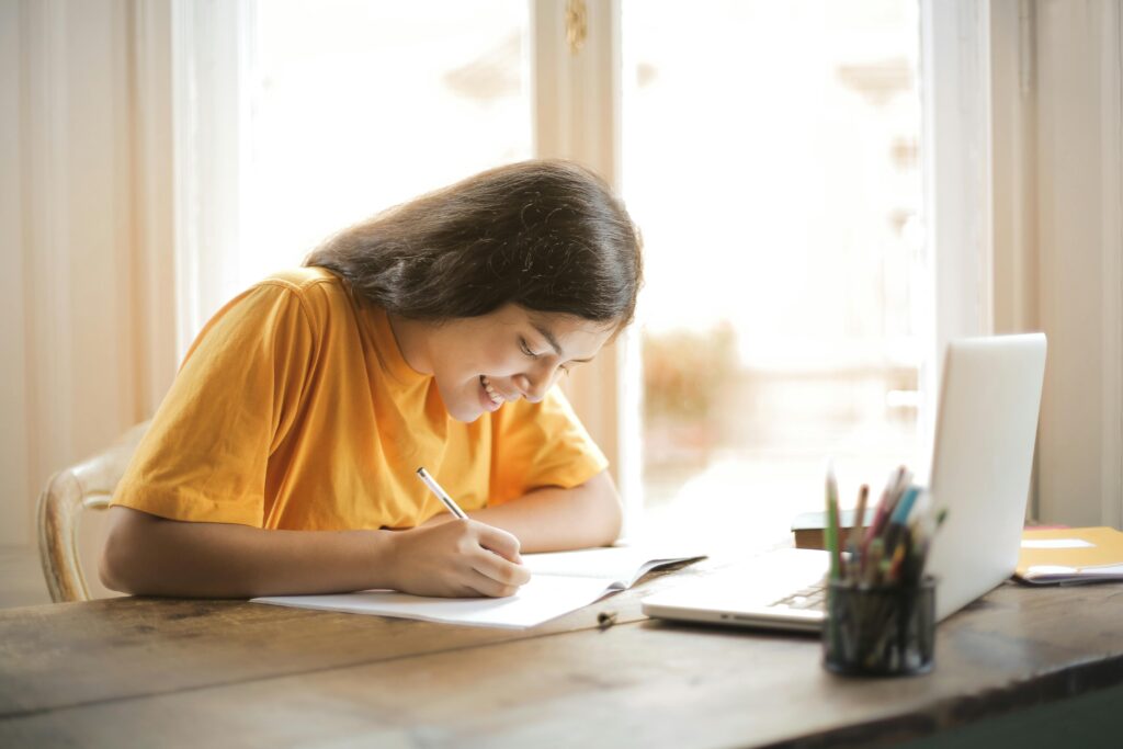 A woman smiling and writing on paper book with her laptop open in front of her.