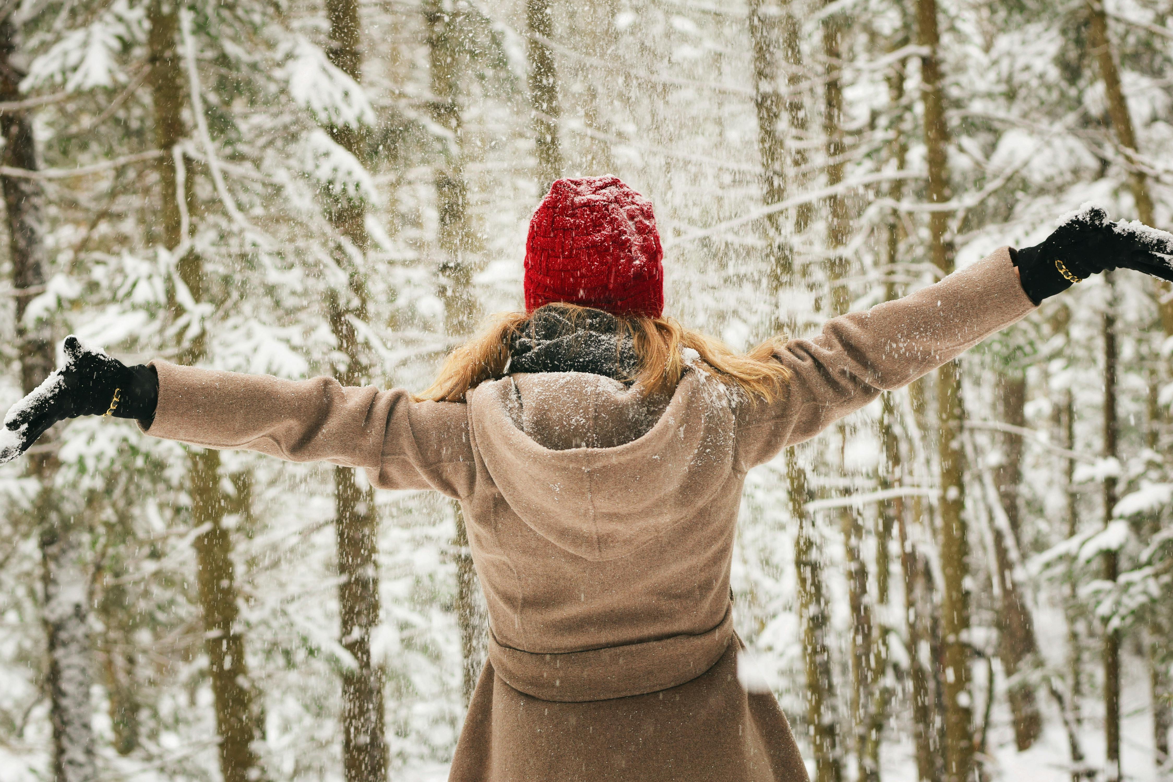 A woman wearing a beige coat and red hat, her back faced towards the camera as she opens her arms wide while looking at the snowy trees