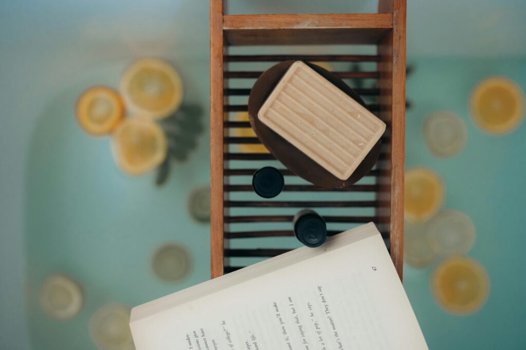 A book and soap on a wooden table above a bath filled with water and lemon slices.